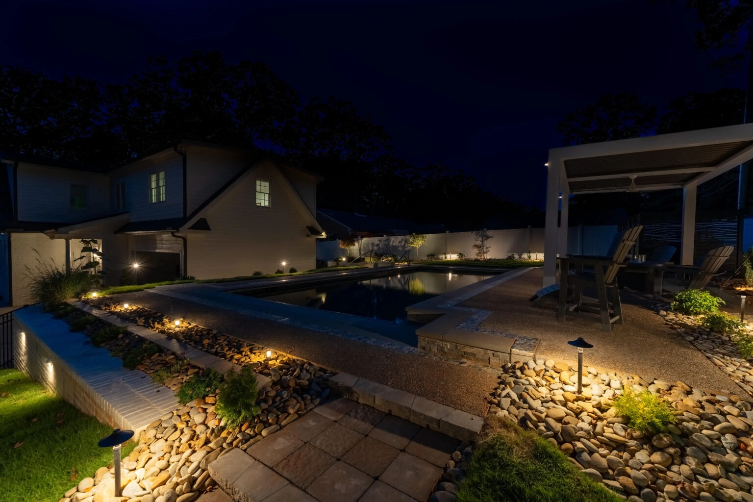 Nighttime view of a backyard pool with landscape lighting, stone beds, a patio, and a covered seating area.